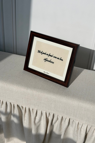 A framed art print with a dark brown wooden frame and cream mat, featuring a handwritten quote in black ink, displayed on a beige fabric-covered table against a gray paneled wall.