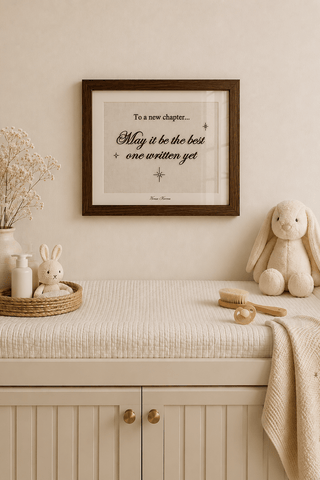 A framed typographic wall art print with a quote hangs above a cream-colored changing table in a styled nursery setting. The room includes a beige bunny plush toy, a small white rabbit figurine, a woven tray with a vase of dried flowers, and a soft knit throw blanket.